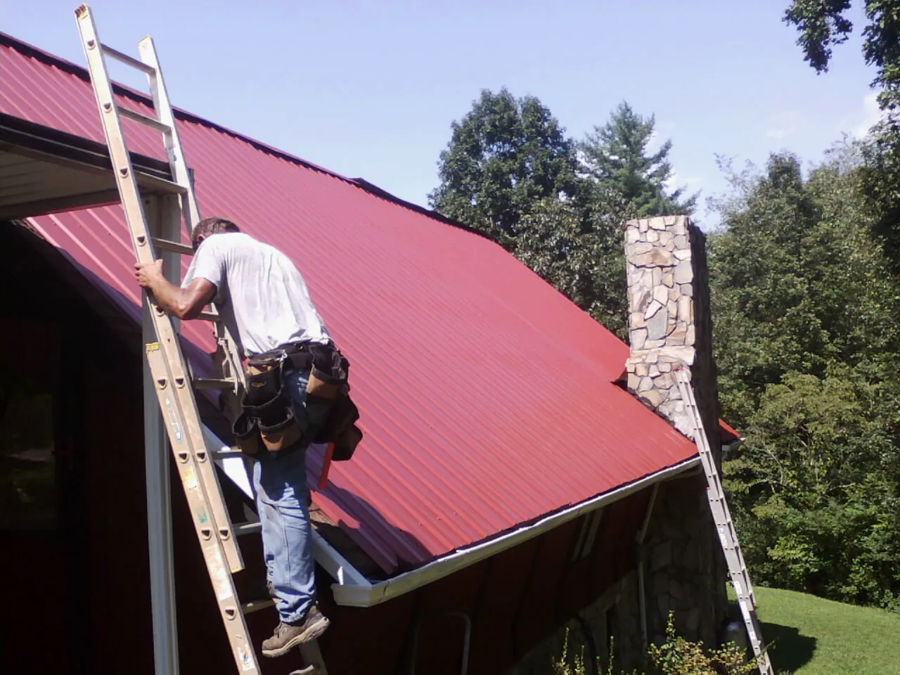 Roofer on a ladder beside a red metal roof and stone chimney — Soffit & Fascia Repair by Roofing Brothers in Landrum